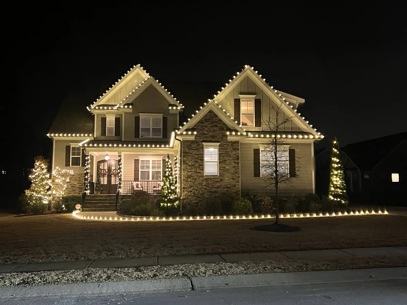 Two-story craftsman home with warm white C9 Christmas lights on every roofline and gable, lit porch columns, wrapped trees and lit landscape bed edging at night