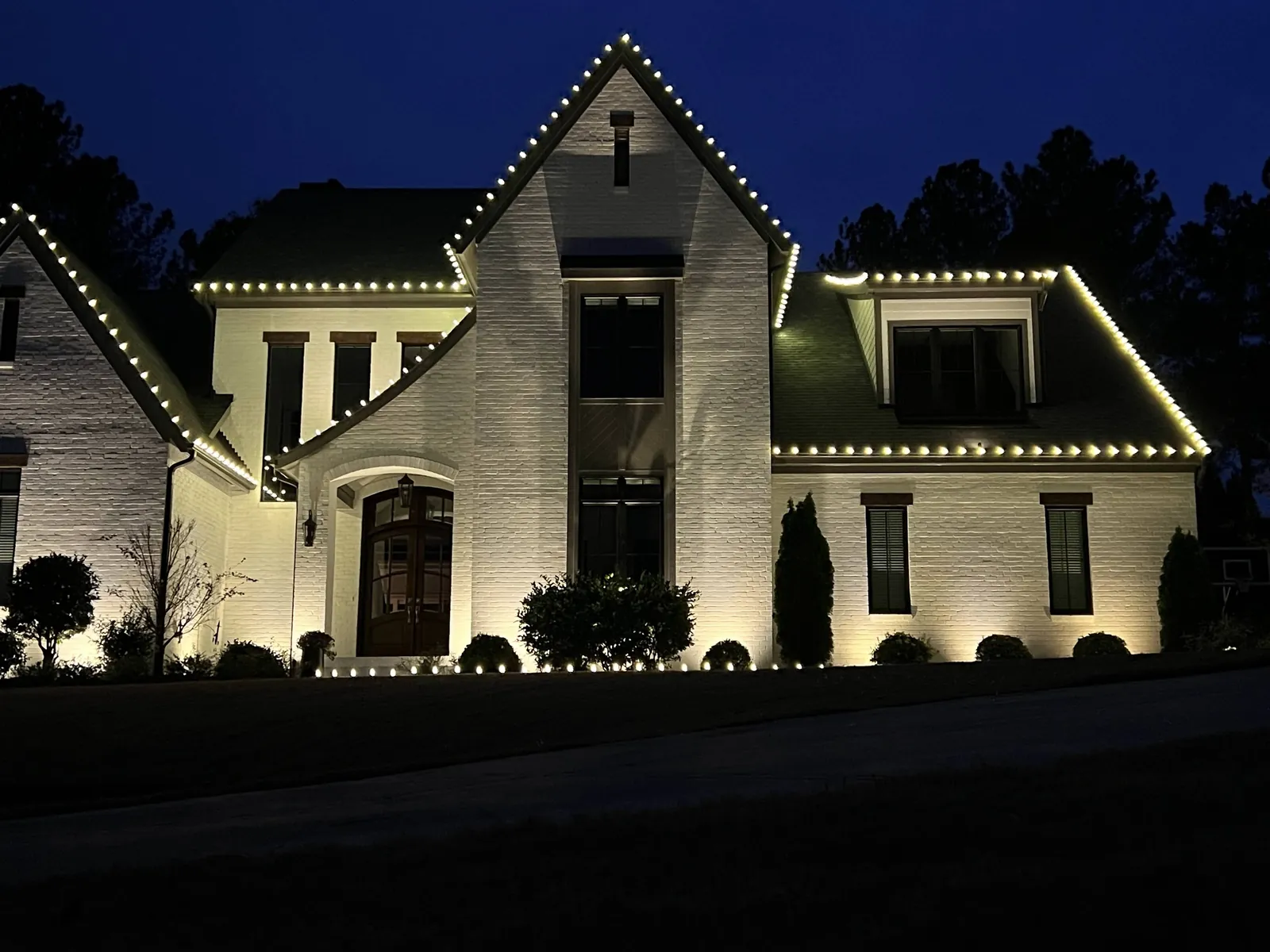 White brick home at dusk with warm white Christmas lights tracing the tall center gable, dormer and eaves