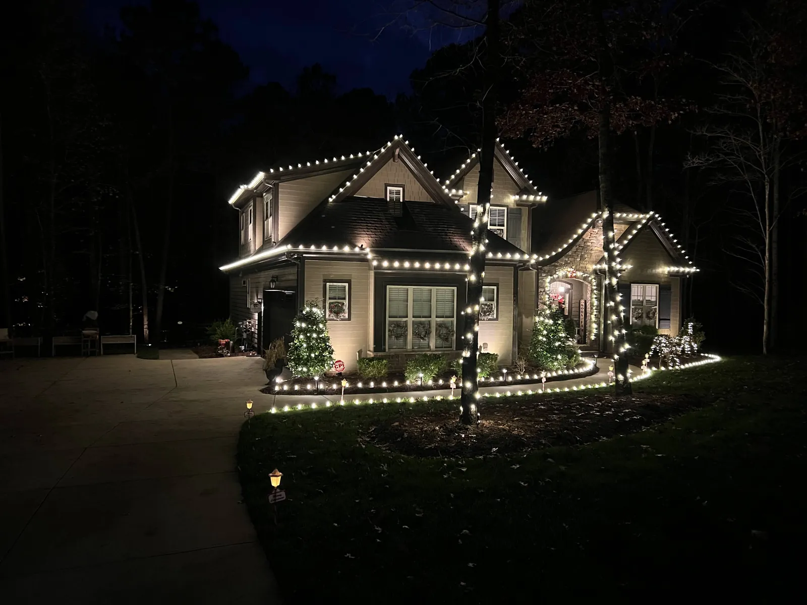 Home at night with warm white Christmas lights on the roofline, wrapped tree trunks, lit shrubs and lights lining the curved walkway and beds