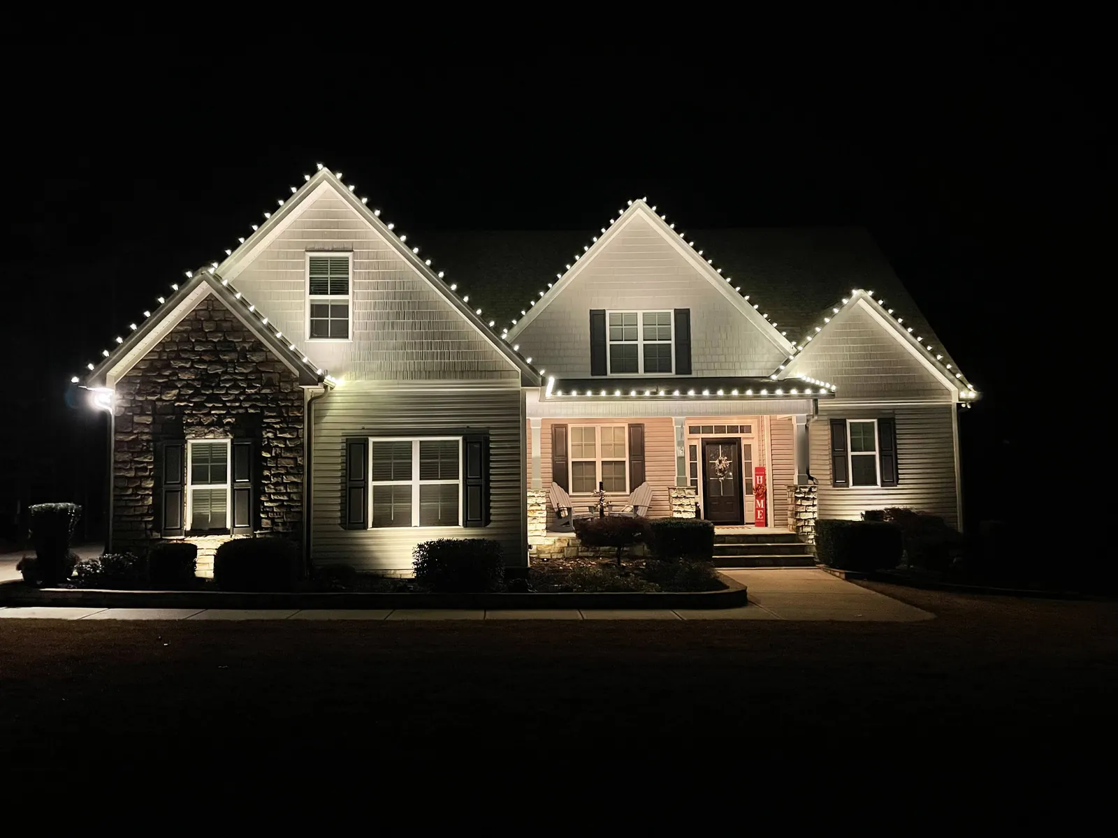 Craftsman-style home with stone accent and three front gables outlined in warm white Christmas lights at night