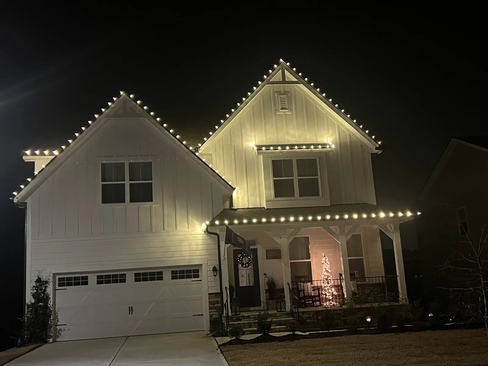 White modern farmhouse with warm white Christmas lights along both steep gables, the window trim and the front porch roofline