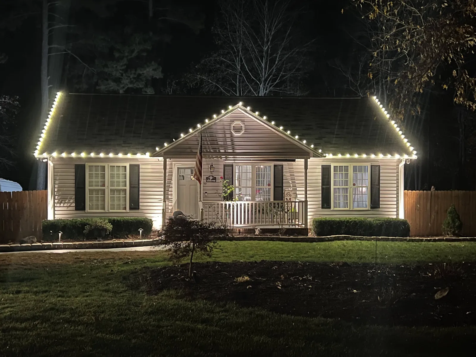 Single-story ranch home with warm white Christmas lights along the roofline, hip corners and front porch gable at night