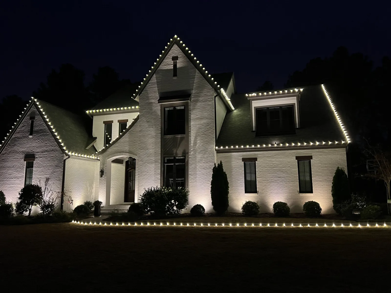 White brick two-story home with warm white Christmas lights outlining steep gables and eaves, plus lights lining the front landscape bed